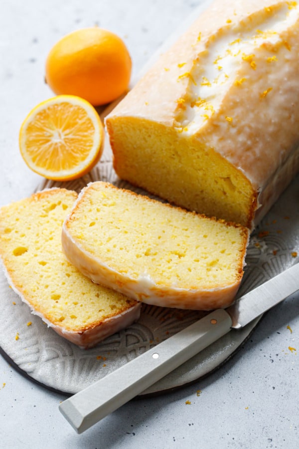 Two cut slices of Meyer Lemon Olive Oil Loaf Cake, showing the tender, moist crumb on the inside and crackly sugar glaze on the outside, with lemons and a knife on a ceramic plate.