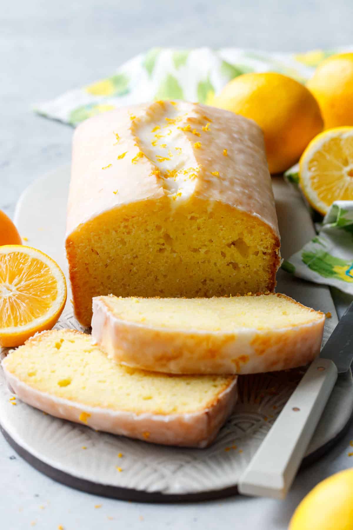 Meyer Lemon Olive Oil Loaf Cake with two slices resting on an oval ceramic plate, with knife, whole and half lemons, and a green and yellow napkin.