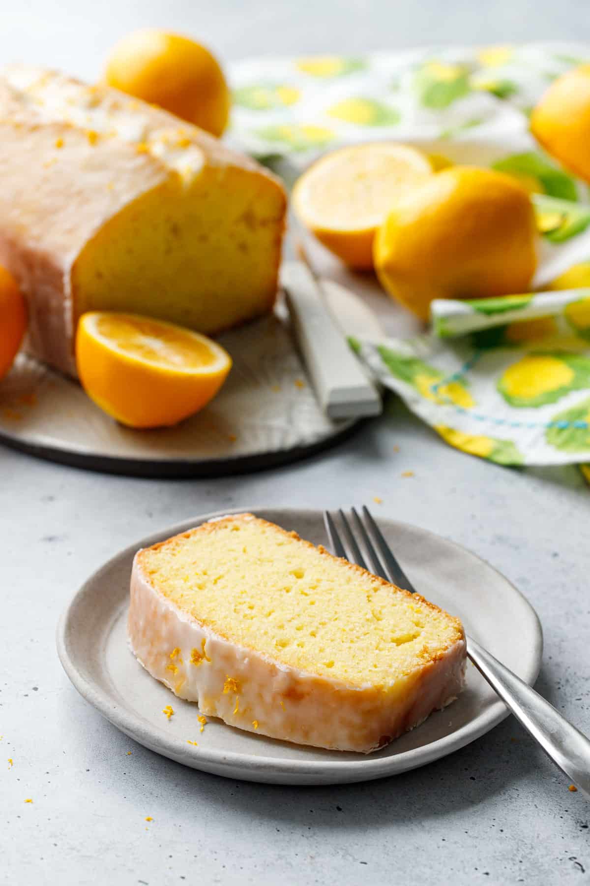 Plate with slice of Meyer Lemon Olive Oil Loaf Cake and a fork, whole loaf, napkin and more lemons in the background.