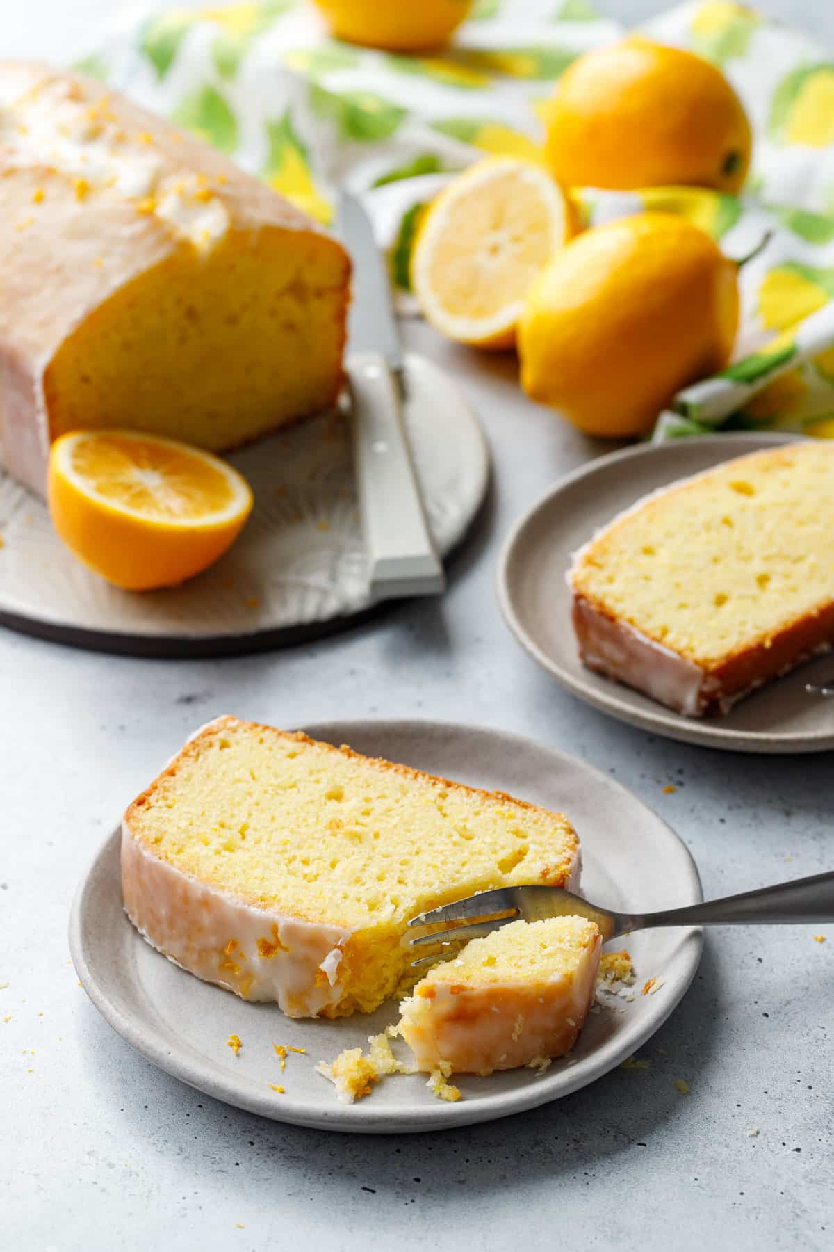 Slice of Meyer Lemon Olive Oil Loaf Cake on a plate with a forkful taken out of it, another slice, whole loaf, and lemons in the background.