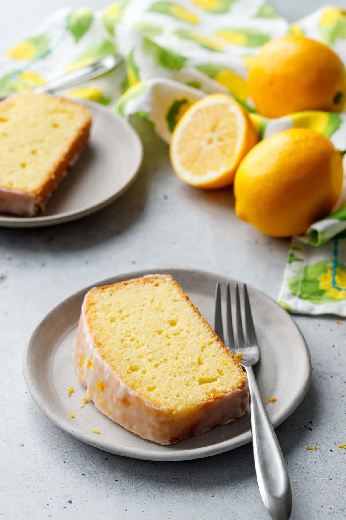 Two ceramic plates with cleanly cut slices of Meyer Lemon Olive Oil Loaf Cake, with green and yellow napkin and Meyer lemons in the background.