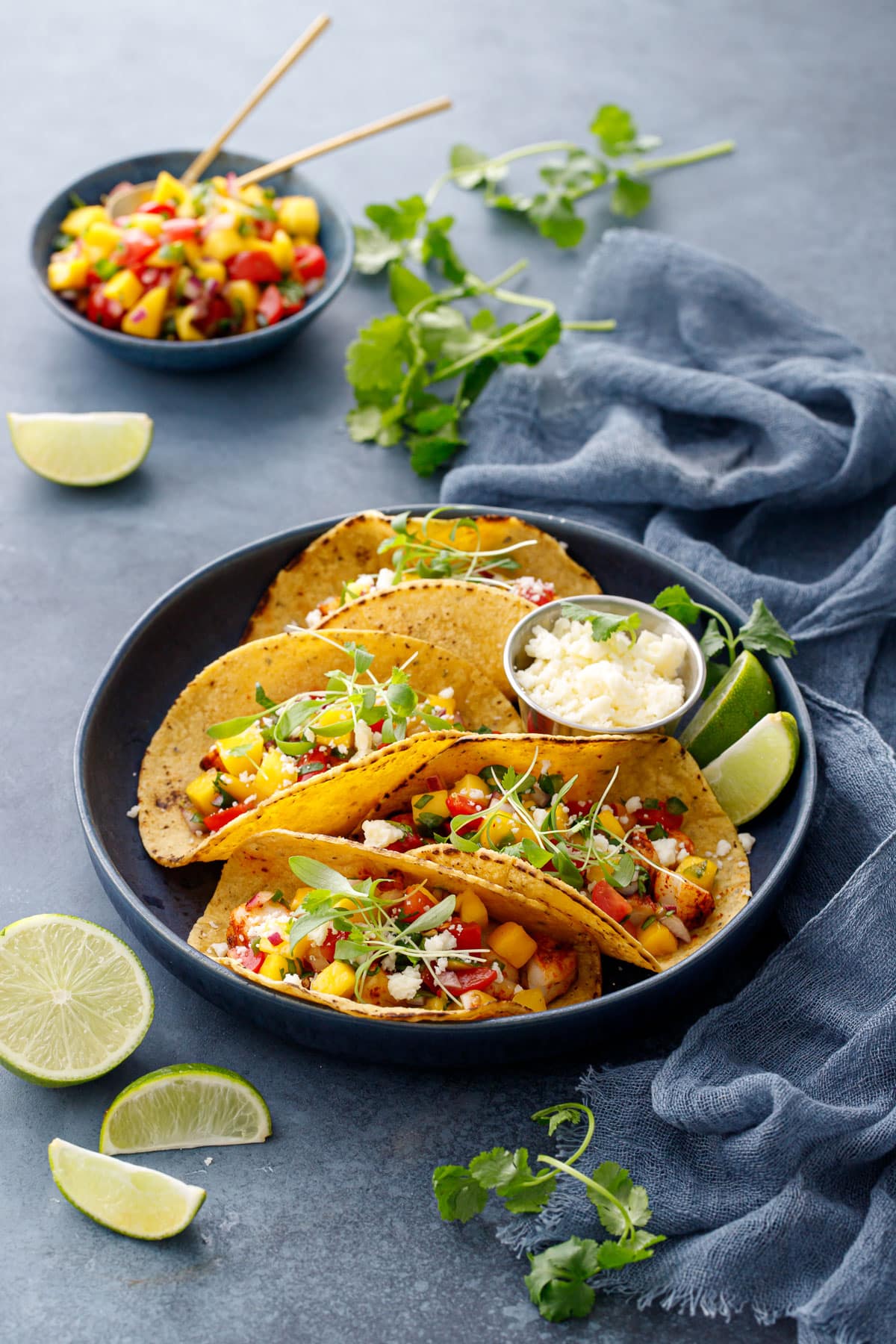 Navy platter of four Shrimp Tacos with yellow corn tortillas and topped with mango salsa and micro cilantro, fresh limes and a bowl of mango salsa on the side.