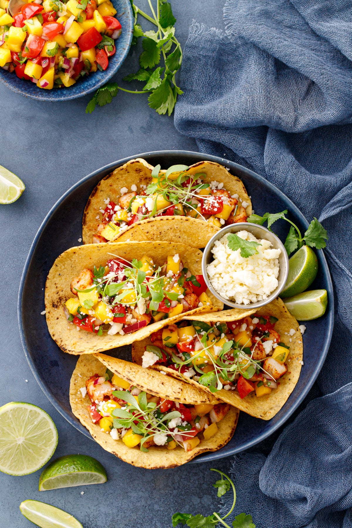 Overhead, navy plate of four shrimp tacos and a bowl of cotija cheese on a blue background, with a bowl of mango salsa and fresh limes and cilantro.