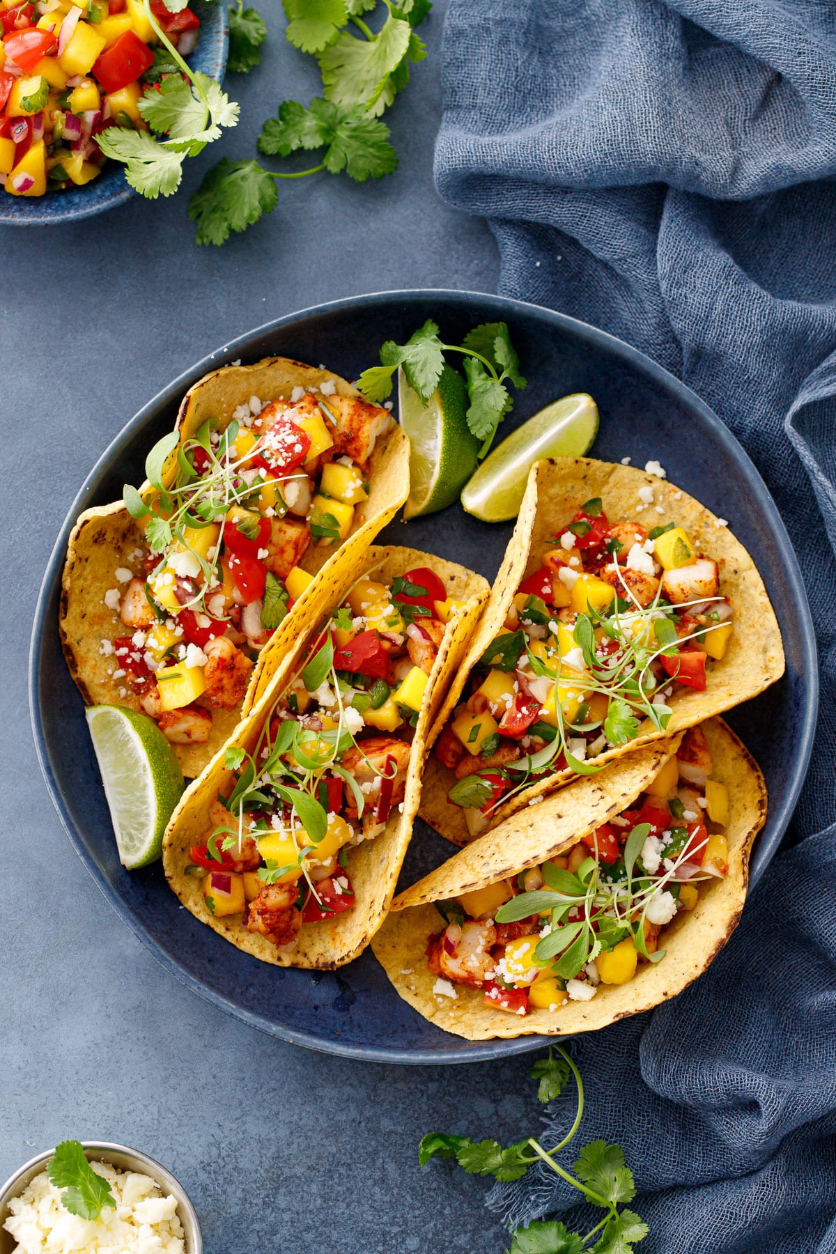 Overhead, dish of four shrimp tacos on a blue background, with a bowl of mango salsa and fresh cilantro scattered around.