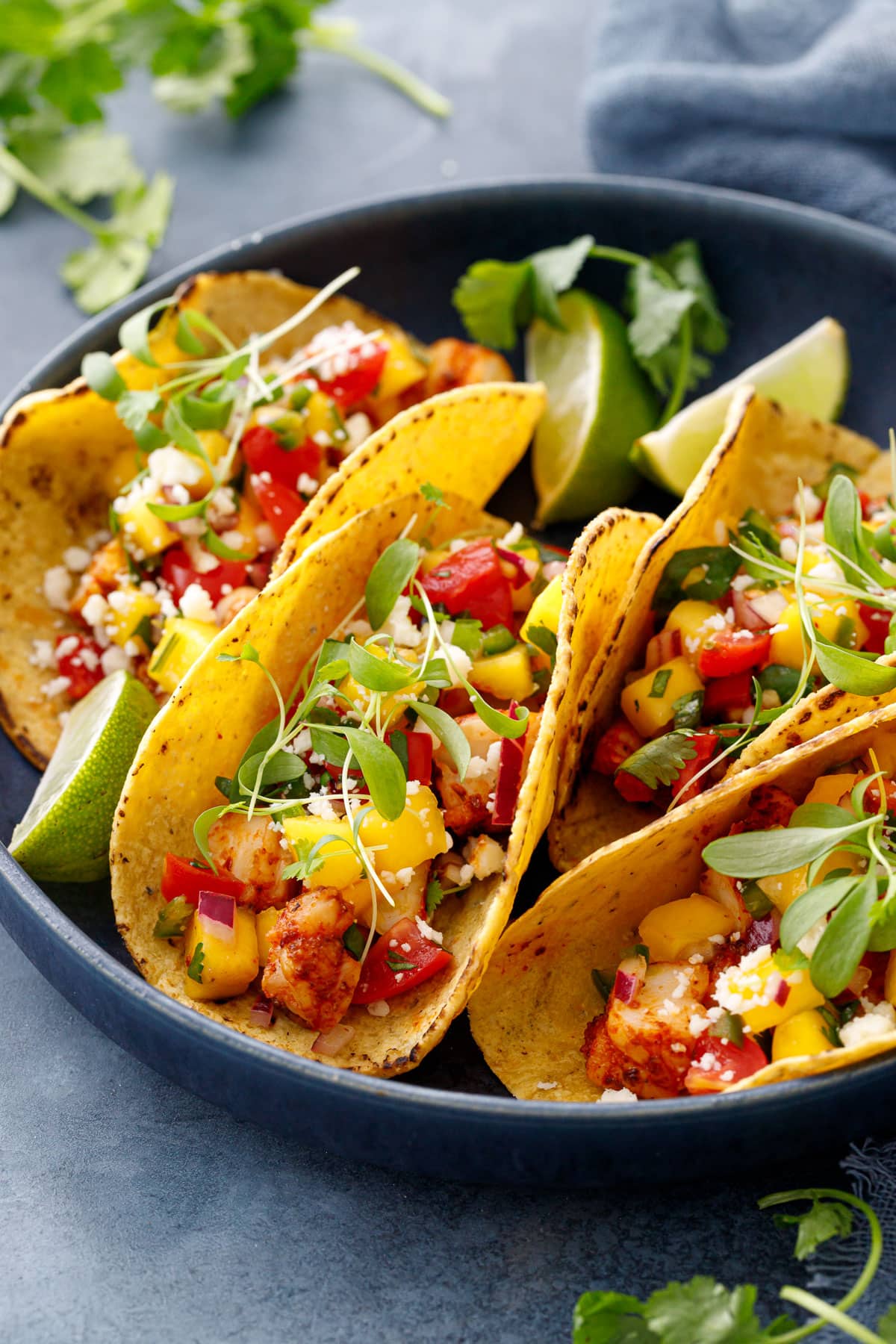 Closeup, shrimp tacos with yellow corn tortillas, topped with a colorful mango salsa, cotija cheese, and micro cilantro with wedges of fresh lime on the side.