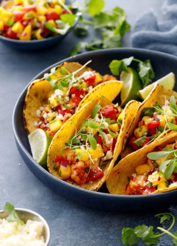 Four shrimp tacos nestled in a navy blue bowl, with a colorful mango salsa and crumbled cotija cheese and micro cilantro on top.