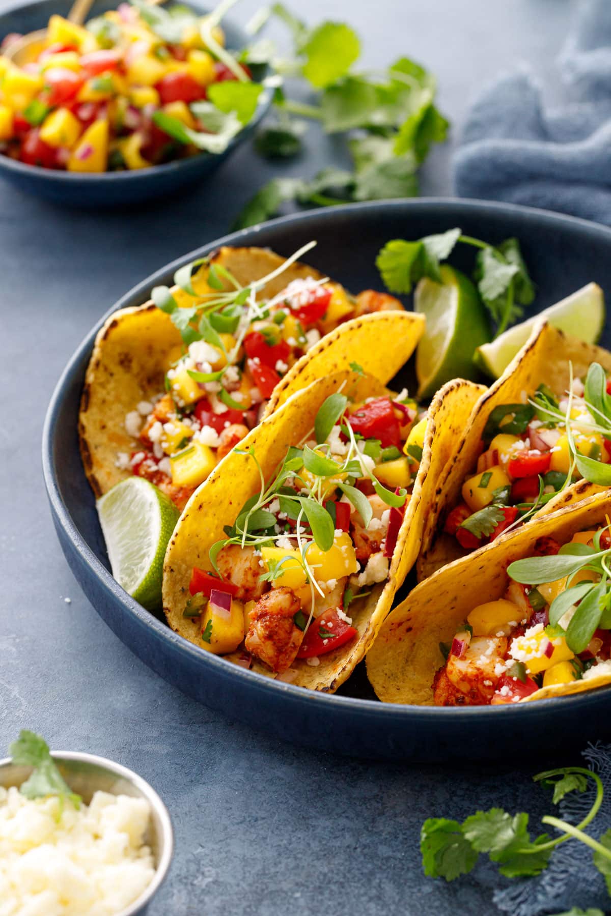 Four shrimp tacos nestled in a navy blue bowl, with a colorful mango salsa and crumbled cotija cheese and micro cilantro on top.