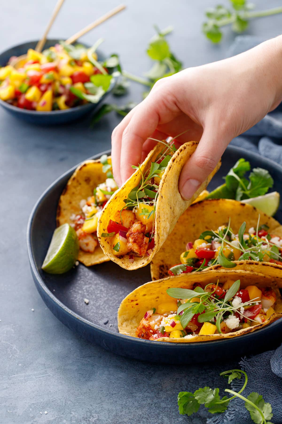 Hand lifting a single shrimp taco out of a navy blue bowl on a blue background, mango salsa in the background.