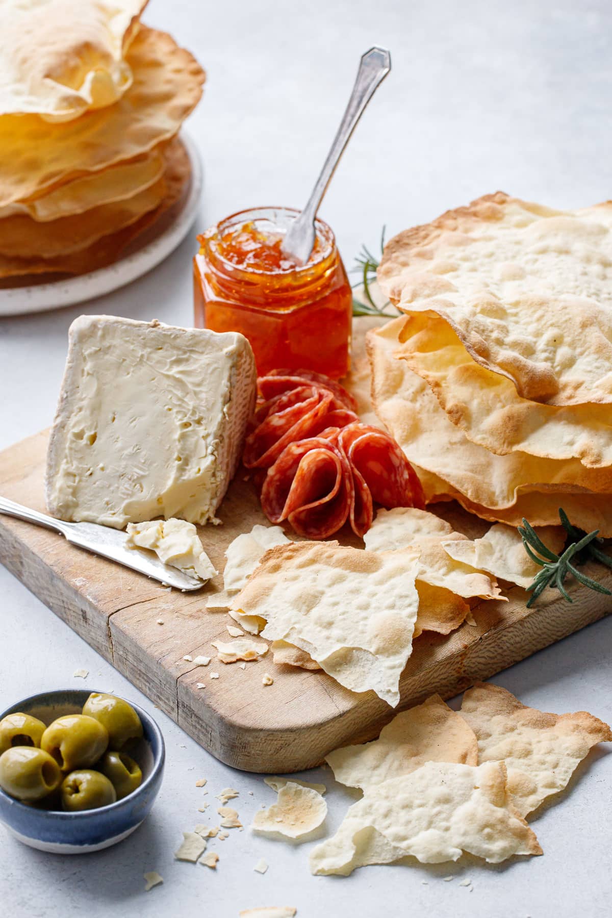 Bread board with Sourdough Semolina Parchment Crackers, some broken into pieces to show the thinness, with soft cheese, salami, pepper jelly and olives.