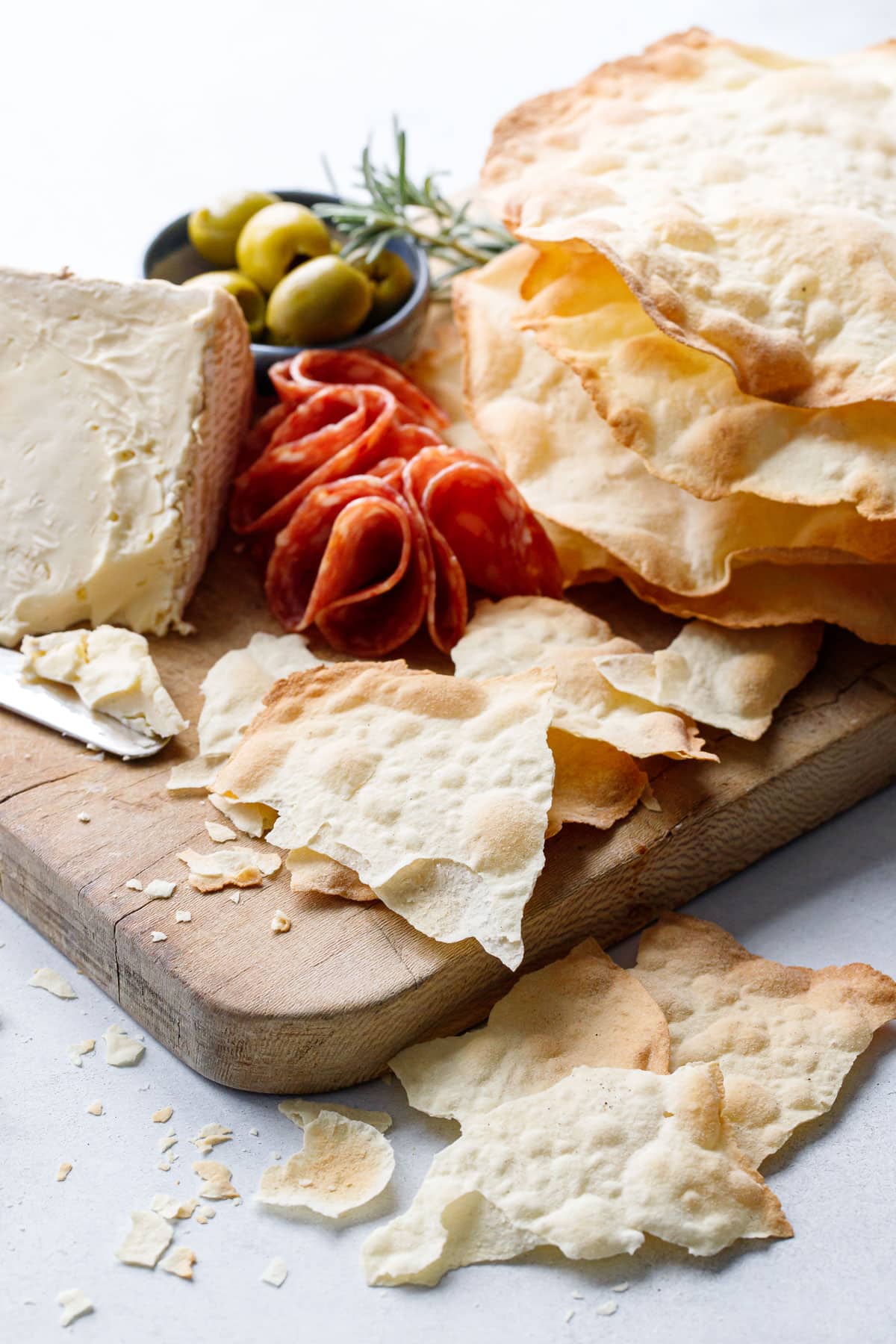 Sourdough Semolina Parchment Crackers on a charcuterie board, some broken into pieces to show the thinness, with soft cheese, salami, pepper jelly and olives.