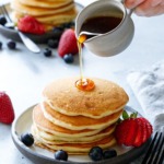 Pouring maple syrup on a tall stack of Olive Oil Pancakes, another plate in the background.