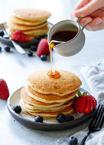 Pouring maple syrup on a tall stack of Olive Oil Pancakes, another plate in the background.