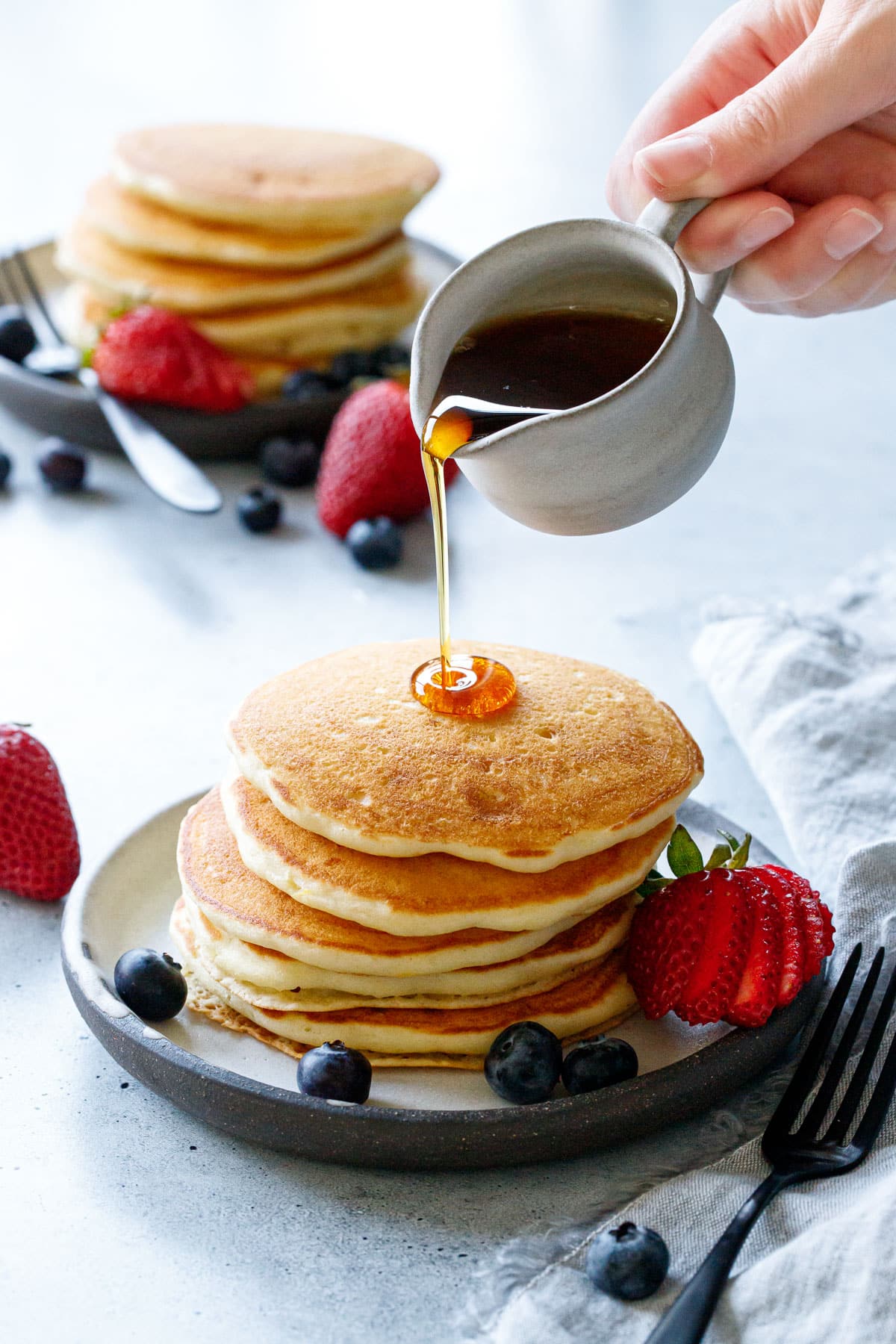 Pouring maple syrup on a tall stack of Olive Oil Pancakes, another plate in the background.