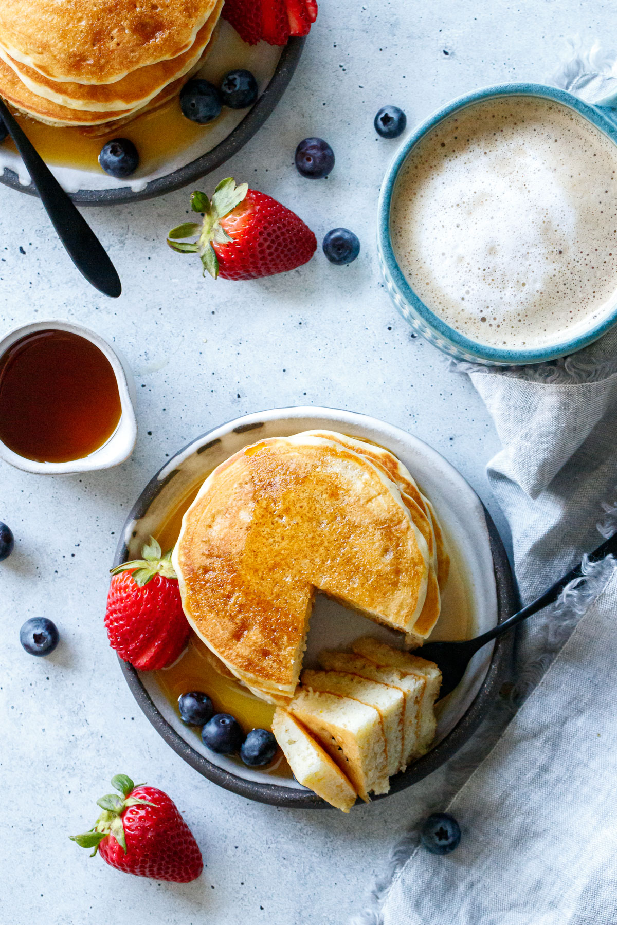 Overhead, two plates of Olive Oil Pancakes, one with a wedge cut out to show texture, plus fruit, cup of coffee, and little pitcher of maple syrup on the side.