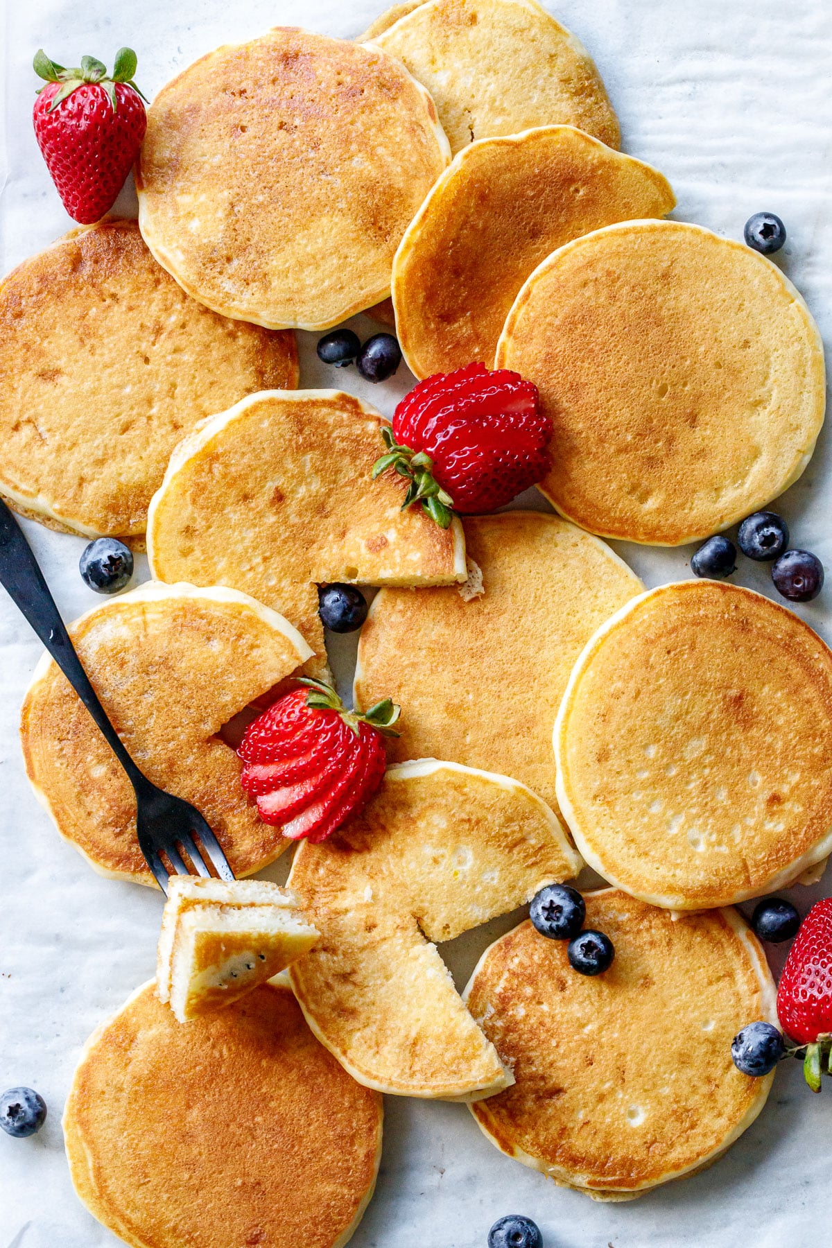 Overhead, scattered arrangement of Olive Oil Pancakes, some cut and some whole, with blueberries and strawberries.
