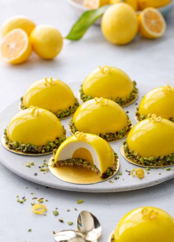 Round plate with bright yellow dome-shaped mousse cakes, one cut to show the layers inside; bowl of lemons in the background.