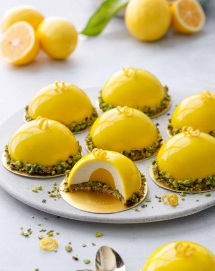 Round plate with bright yellow dome-shaped mousse cakes, one cut to show the layers inside; bowl of lemons in the background.