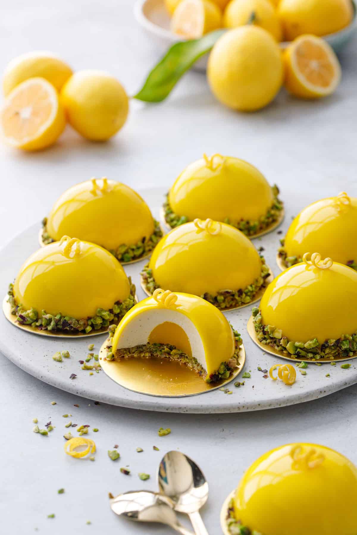 Round plate with bright yellow dome-shaped mousse cakes, one cut to show the layers inside; bowl of lemons in the background.