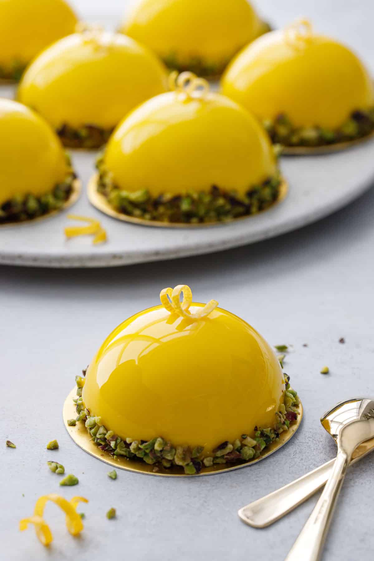 Bright yellow mirror-glazed dome shaped mousse cakes with pistachios around the edges, one cake in front and a plate of more in the background.