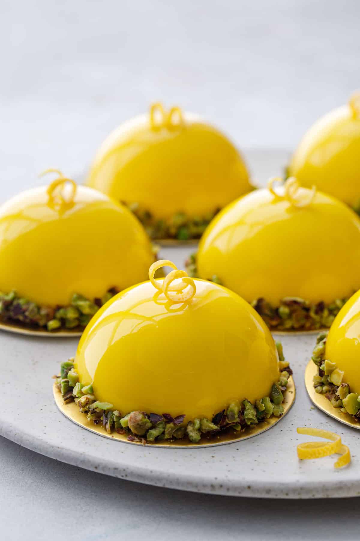 Plate of bright yellow mirror glazed mousse cakes with pistachio edges and a curl of lemon peel on top.