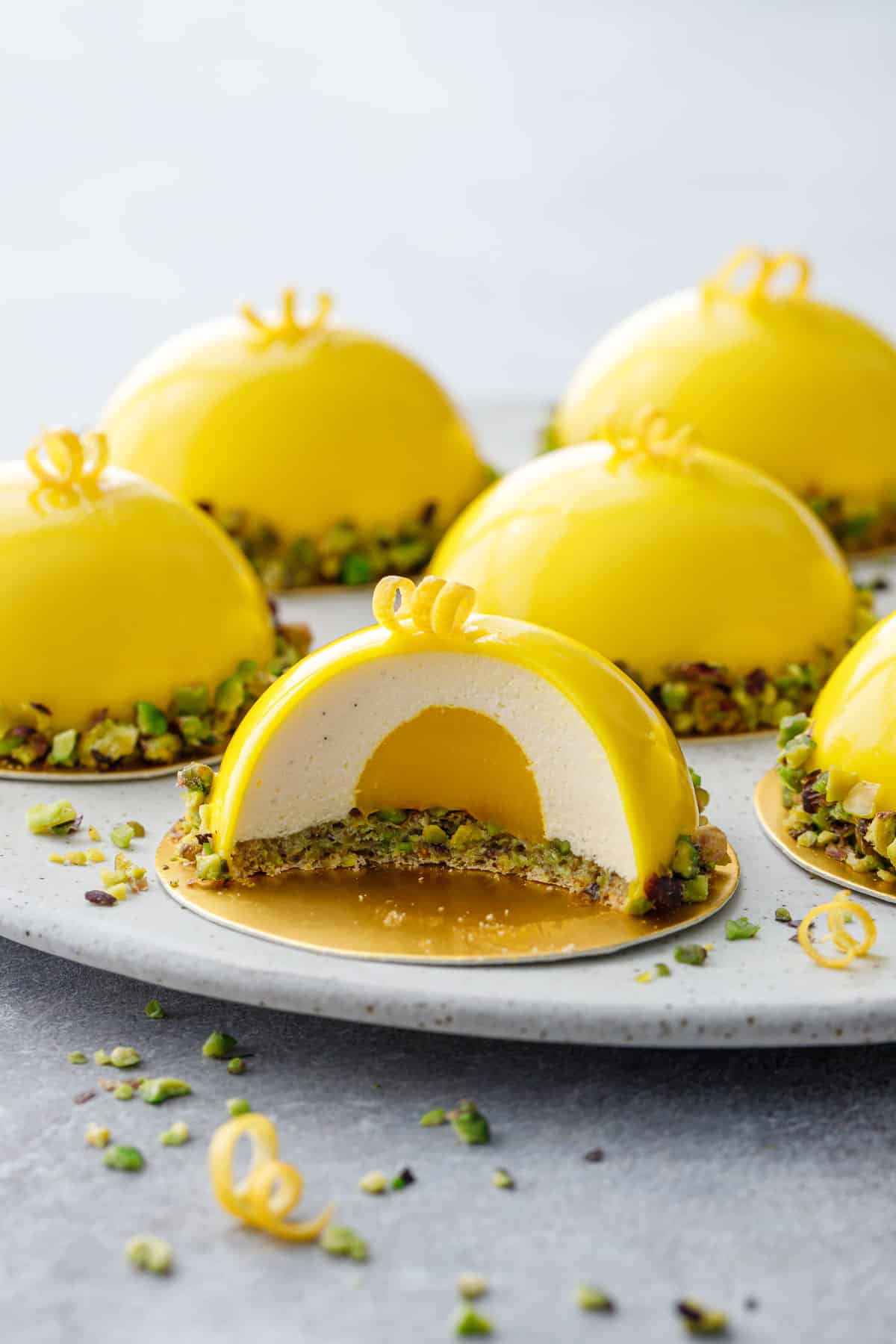 Plate with Pistachio & Meyer Lemon Mousse Cakes with bright yellow mirror glaze, cross section of one cake showing layers inside.