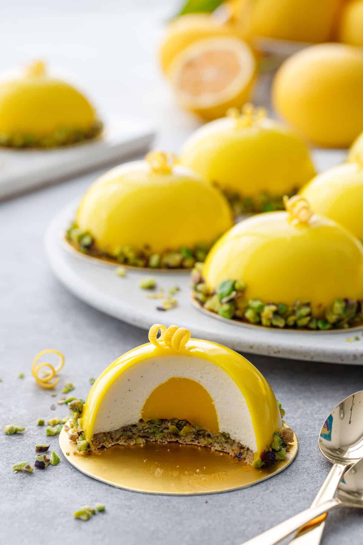 Pistachio & Meyer Lemon Mousse Cake cut into a cross section showing the distinct layers of pistachio dacquoise, lemon curd, lemon mousse, and bright yellow mirror glaze.