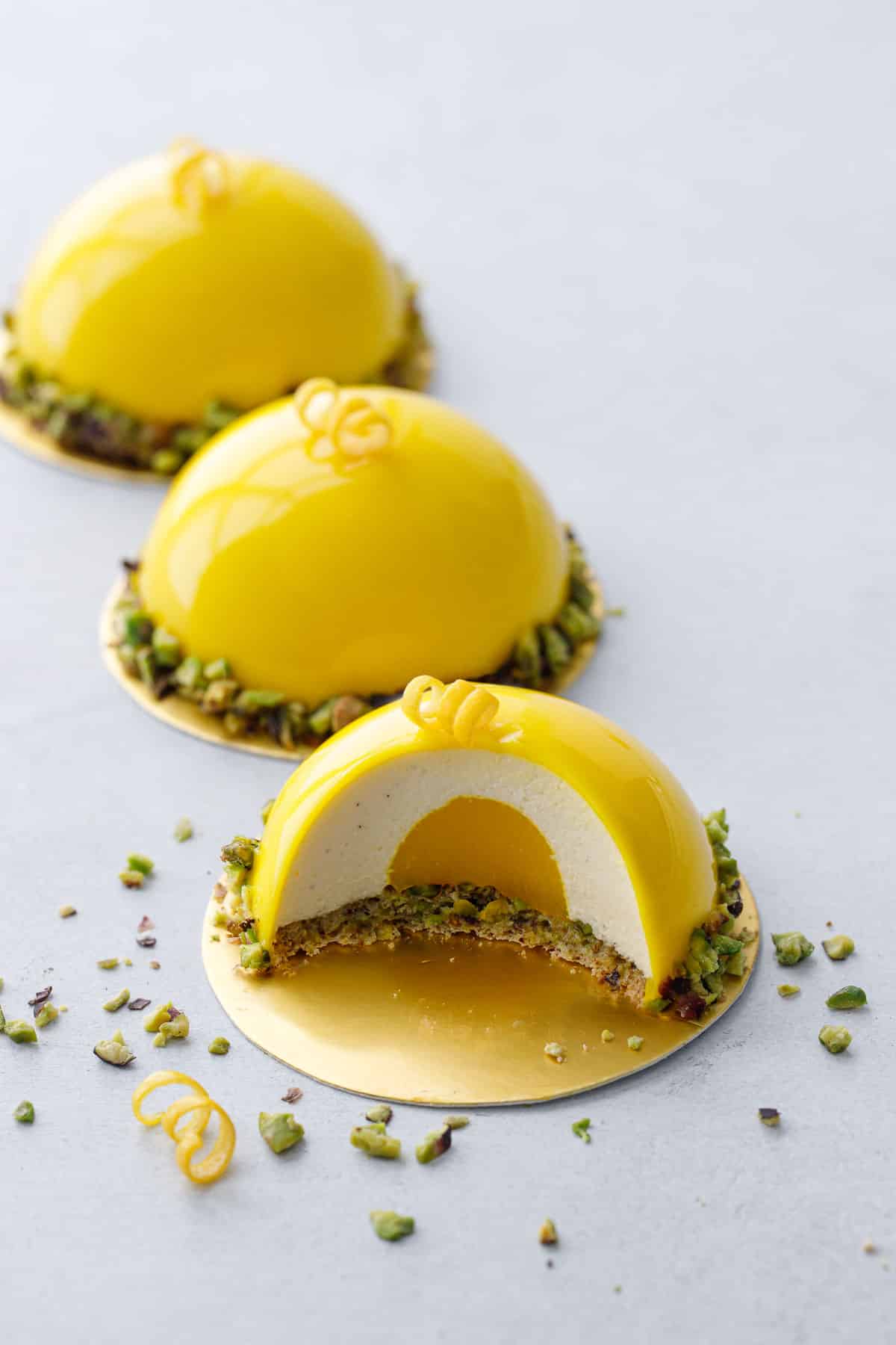 Three Pistachio & Meyer Lemon Mousse Cakes in a row on a gray background, one cut to show the lemon curd center and pistachio dacquoise base.