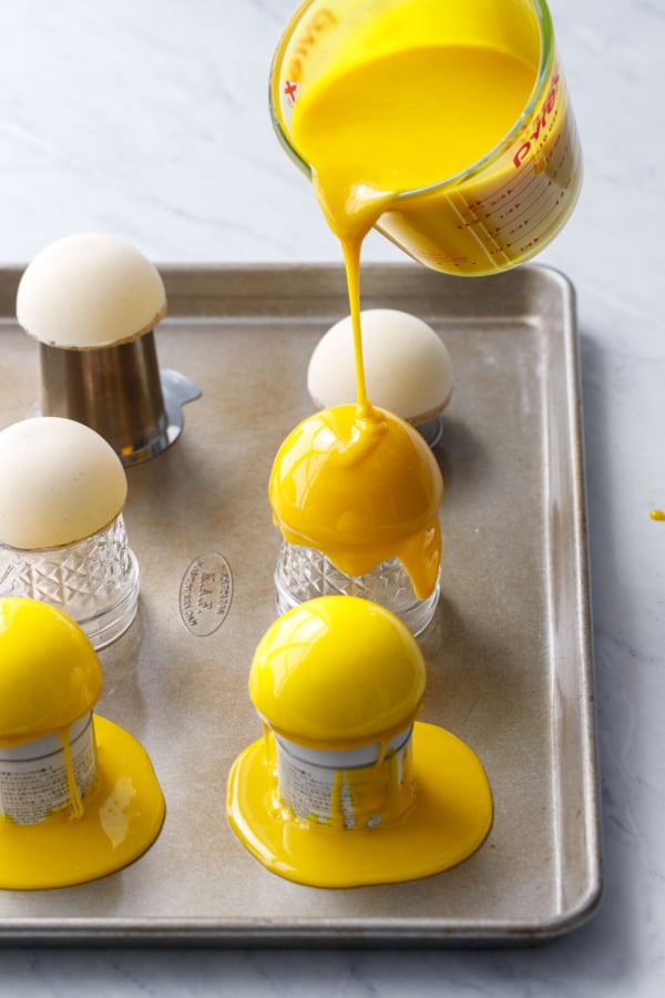 Pouring bright yellow mirror glaze over dome-shaped mousse cakes, allowing the excess to drip off the sides.