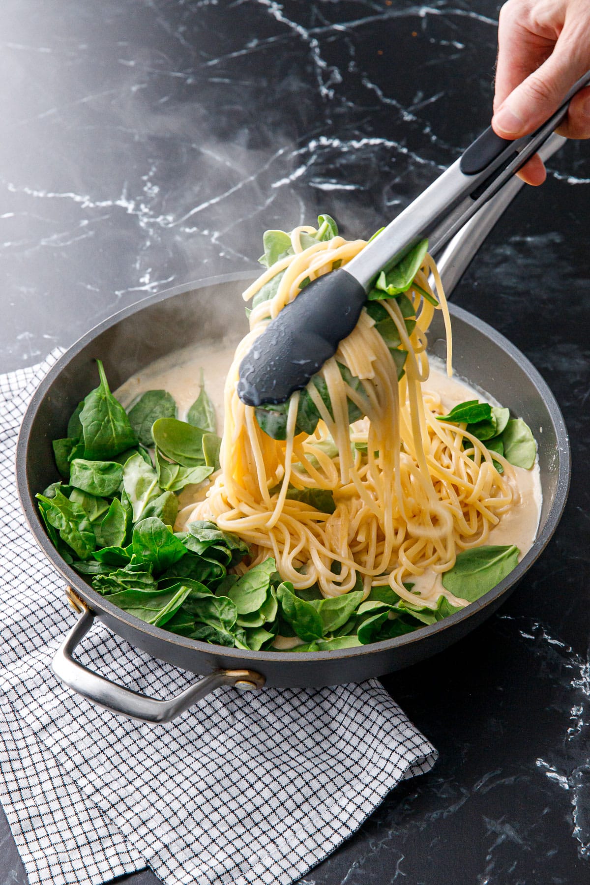 Nonstick skillet with linguine pasta and baby spinach, tongs lifting a scoop of pasta as it is stirred into the creamy sauce.