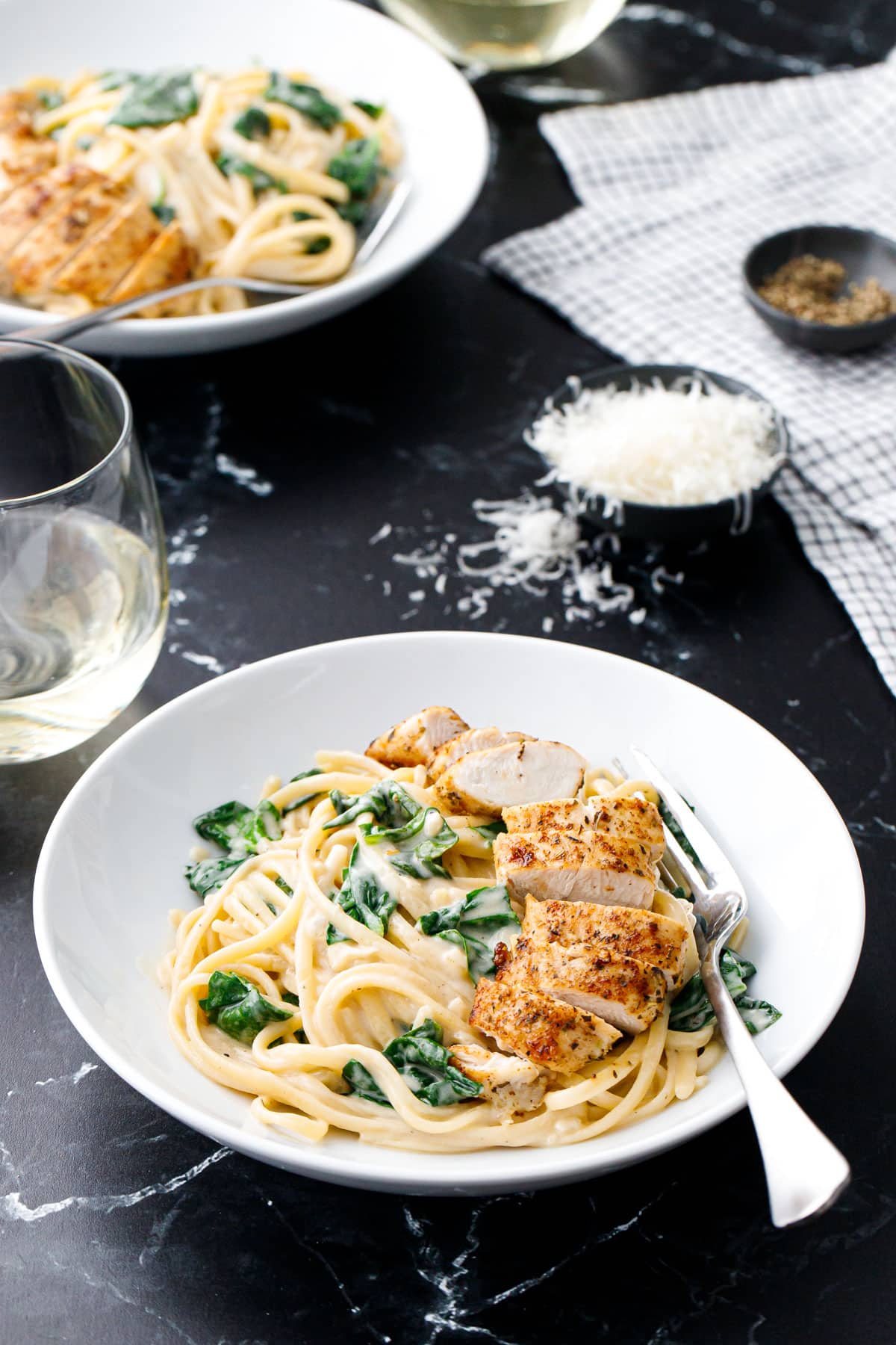 White bowls of Creamy Chicken Florentine Pasta with silver forks, dishes of parmesan cheese and black pepper, plus white wine glasses on the side.