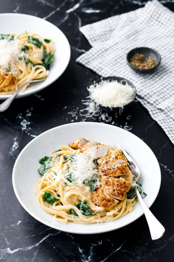 White bowls with Creamy Chicken Florentine Pasta, silver forks, on a black marble background with bowl of cheese and black pepper on the side.