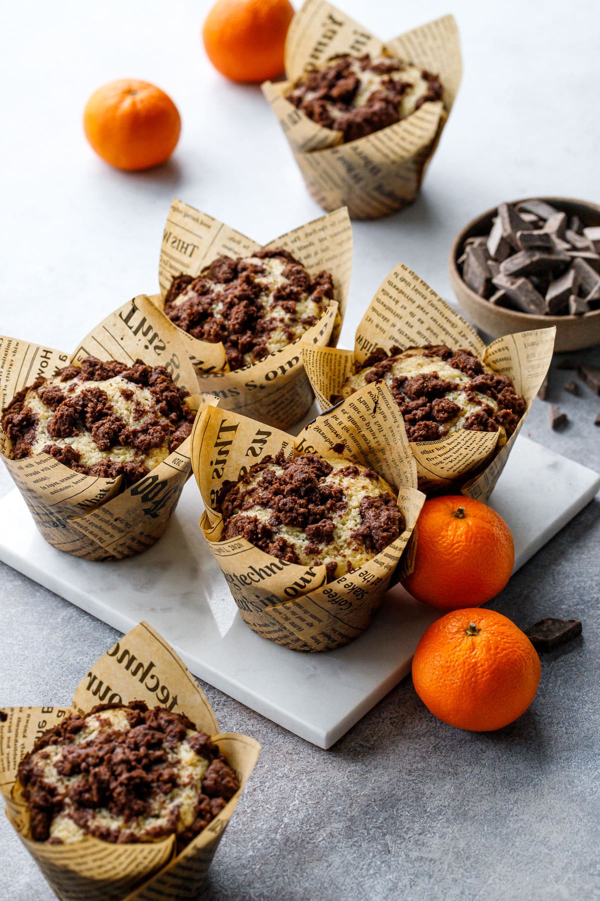 Chocolate Orange Streusel Muffins in natural newsprint tulip wrappers, on a square marble trivet with mandarin oranges on the side.