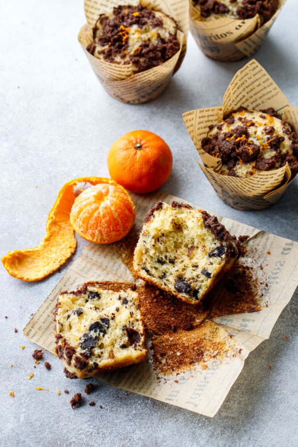 Chocolate Orange Streusel Muffin broken in half to show the texture, with one whole and one peeled orange on the side to indicate the flavor of the muffins.