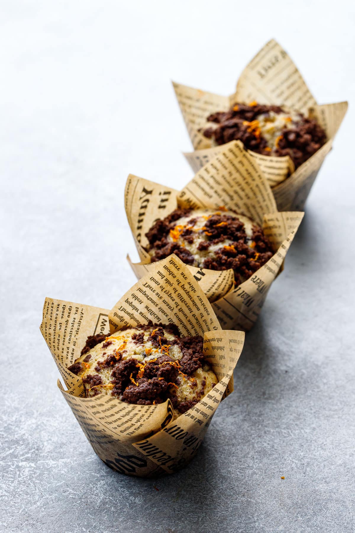 Row of Chocolate Orange Muffins with chocolate streusel on top, on a gray background, baked in newsprint tulip wrappers.