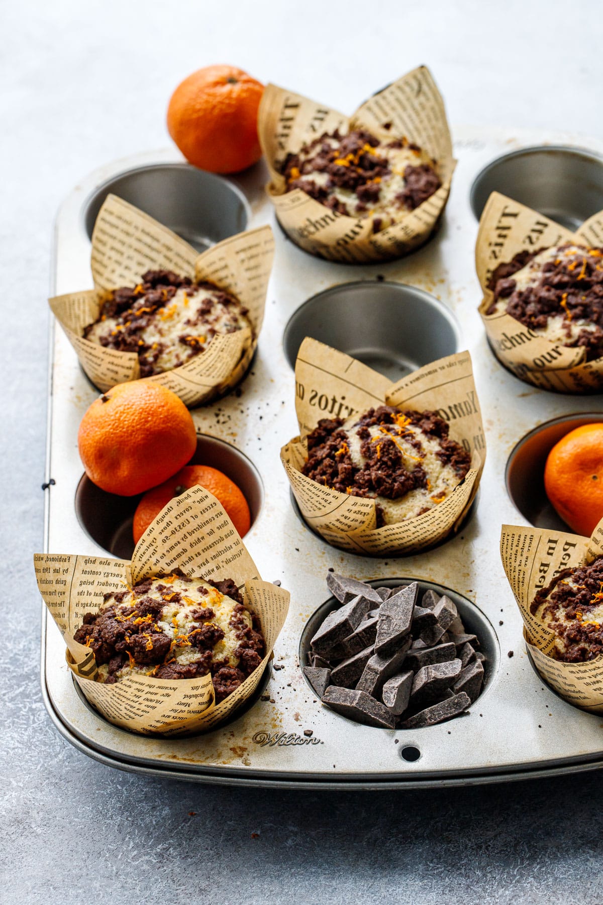 Muffin tin with alternating cups filled with baked muffins in tulip wrappers, some empty cups filled with chocolate chunks and mandarin oranges.