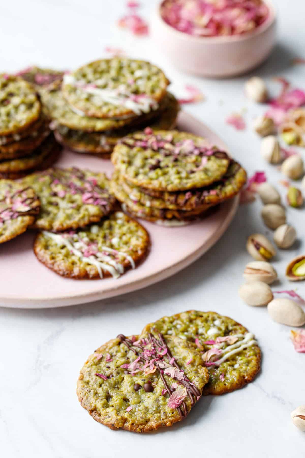 Pistachio Florentine Cookies on a pink plate with scattered pistachios and rose petals around.