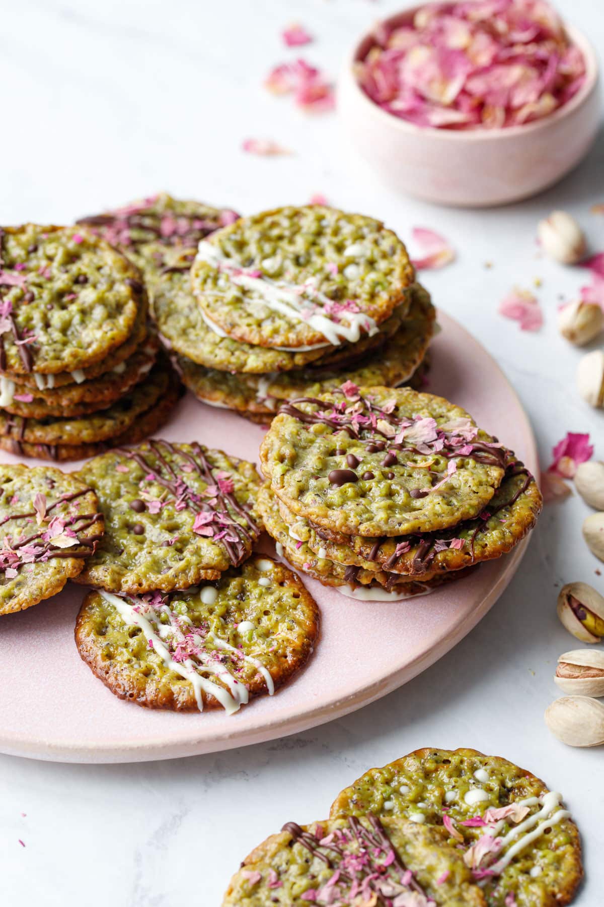 Messy stacks of Pistachio Florentine Cookies on a pink plate with bowl of rose petals and pistachios scattered around.