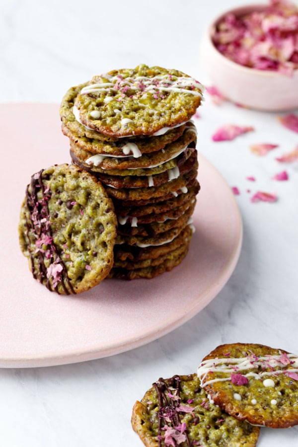 Stack of Pistachio Florentine Cookies on a pink plate, more cookies on the side and a bowl of dried rose petals in the background.