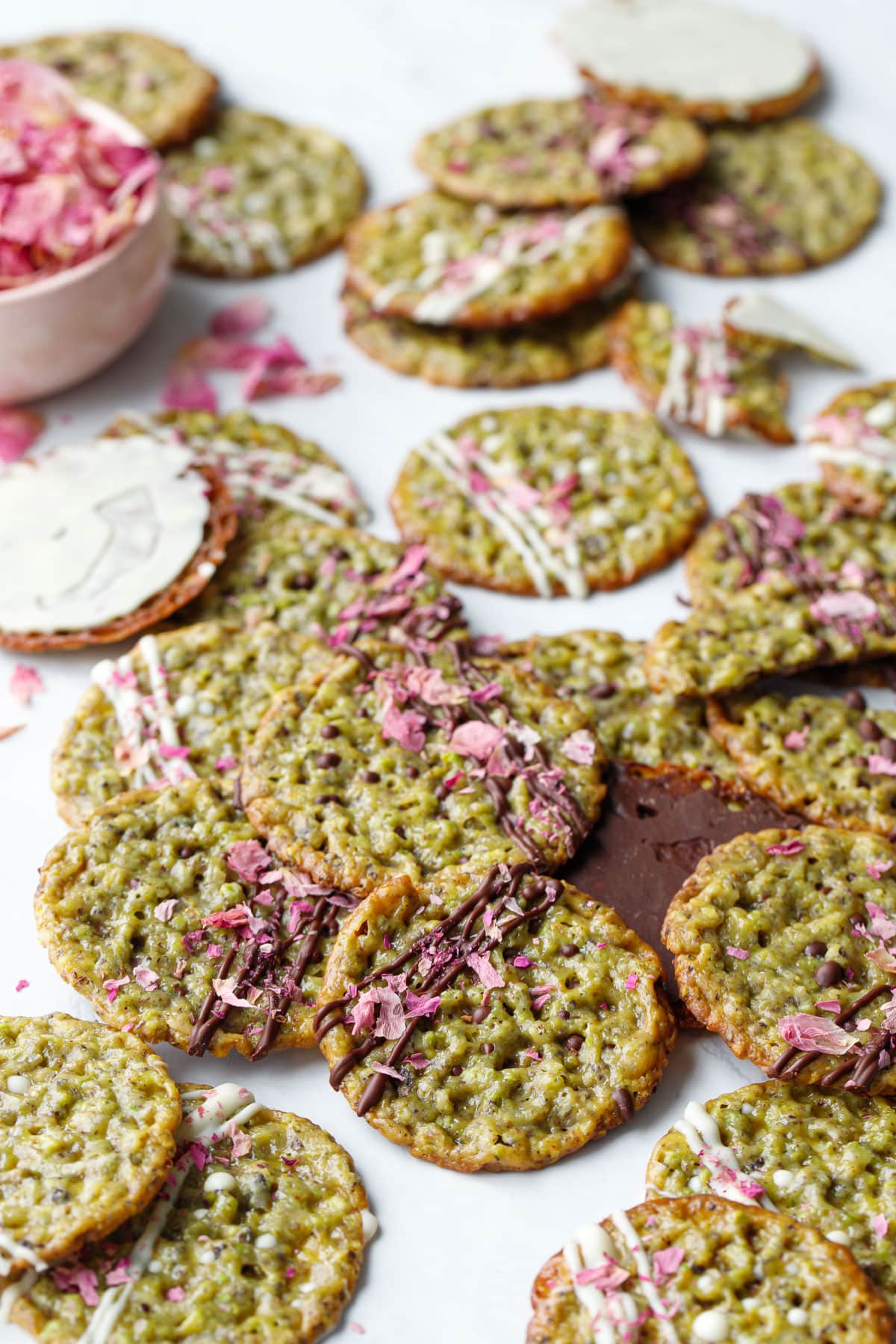 Messy arrangement of Pistachio Florentine Cookies, some with white chocolate drizzles and some with dark, topped with dried rose petals.