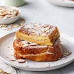 Plate with stack of Marzipan-Stuffed French Toast sprinkled with almonds and dusted with powdered sugar, one piece cut to show the texture of the filling.