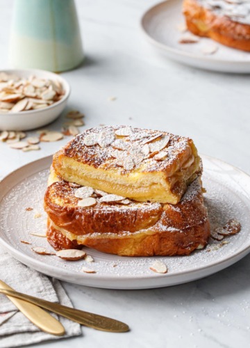 Plate with stack of Marzipan-Stuffed French Toast sprinkled with almonds and dusted with powdered sugar, one piece cut to show the texture of the filling.