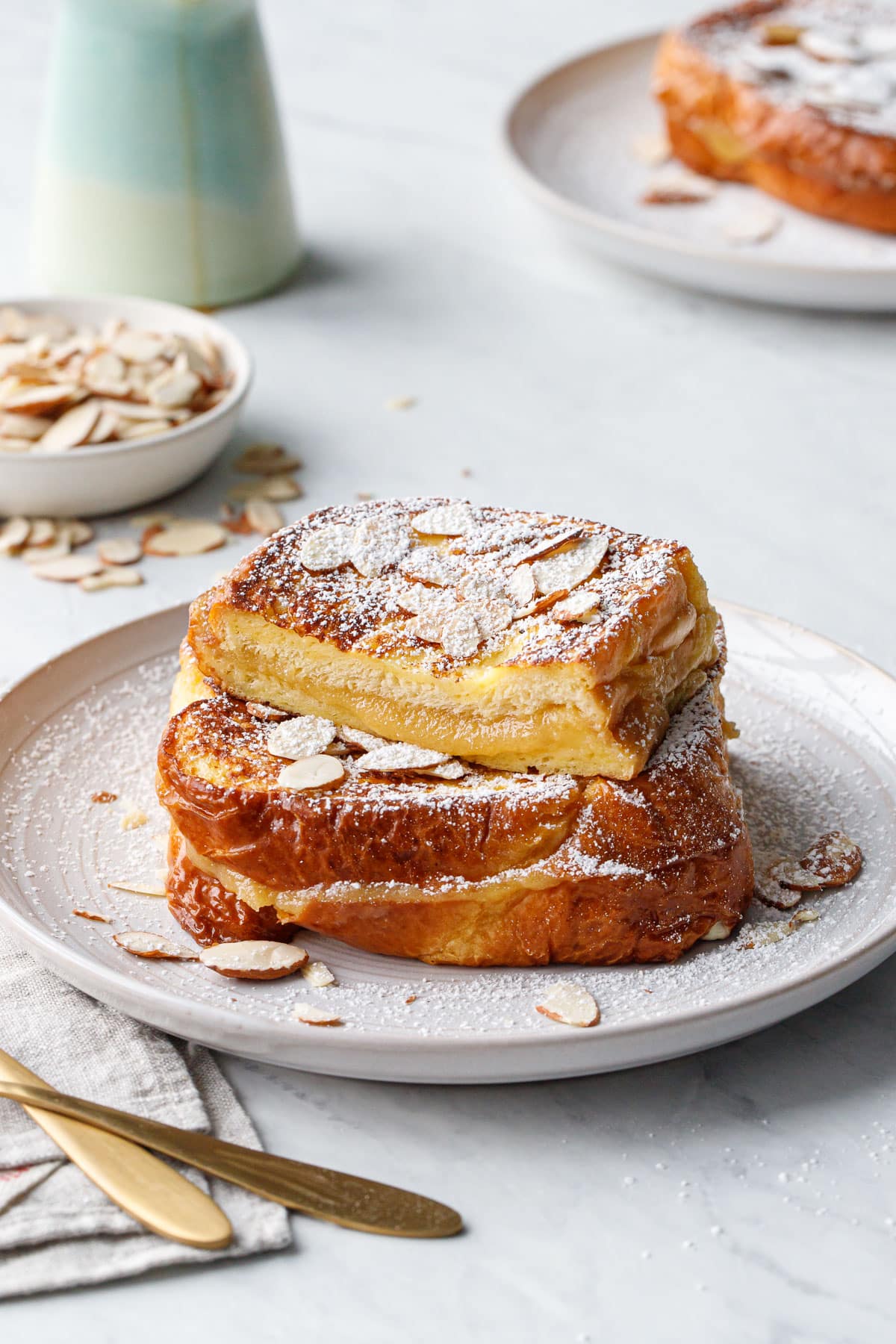 Plate with stack of Marzipan-Stuffed French Toast sprinkled with almonds and dusted with powdered sugar, one piece cut to show the texture of the filling.