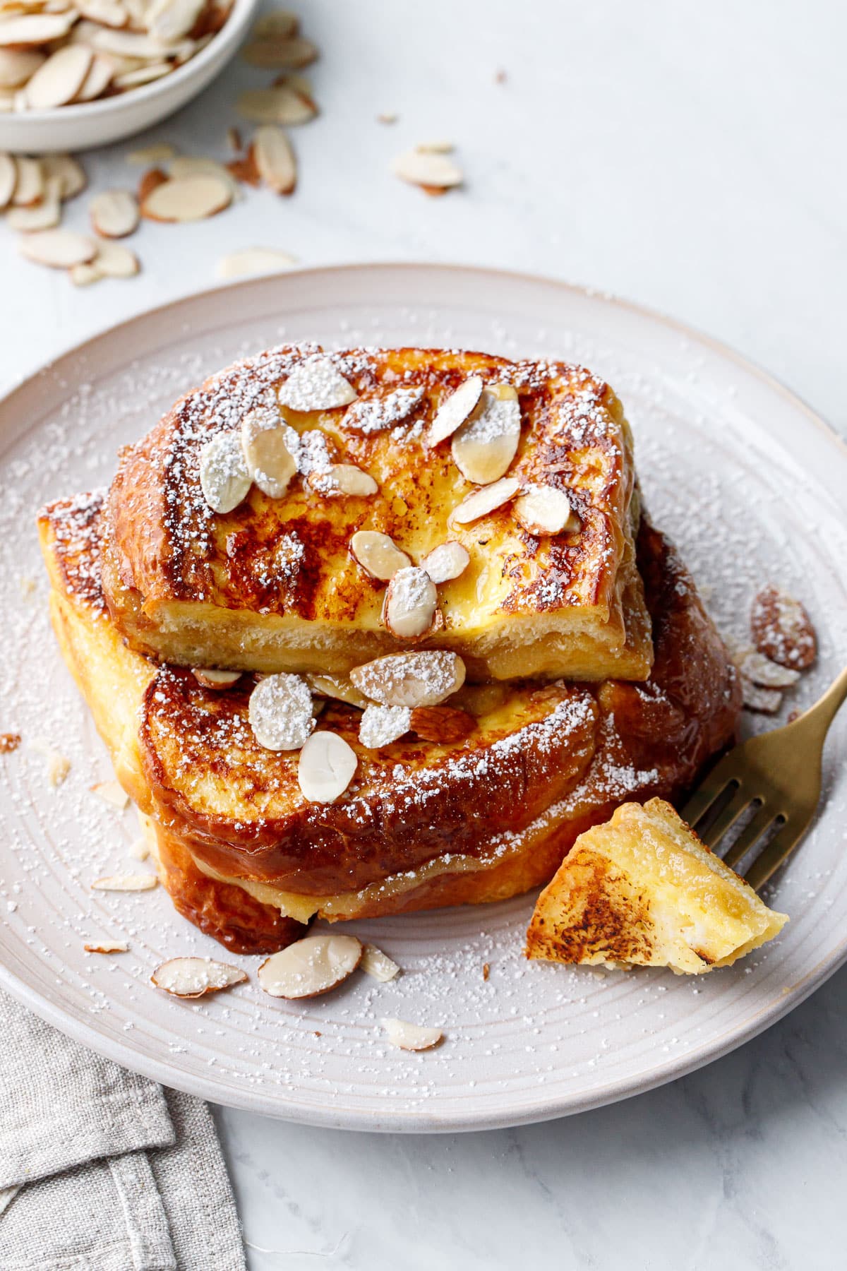 Golden brown Marzipan-Stuffed French Toast topped with sliced almonds and powdered sugar, a cross section bite on a fork that shows the gooey filling.