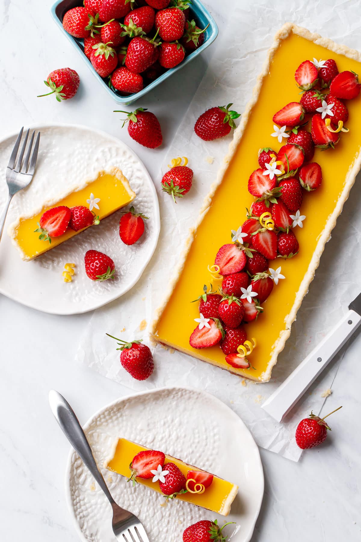 Overhead, plates with slices of Strawberry Meyer Lemon Tart, plus the whole rectangular tart and a basket of strawberries on the side.
