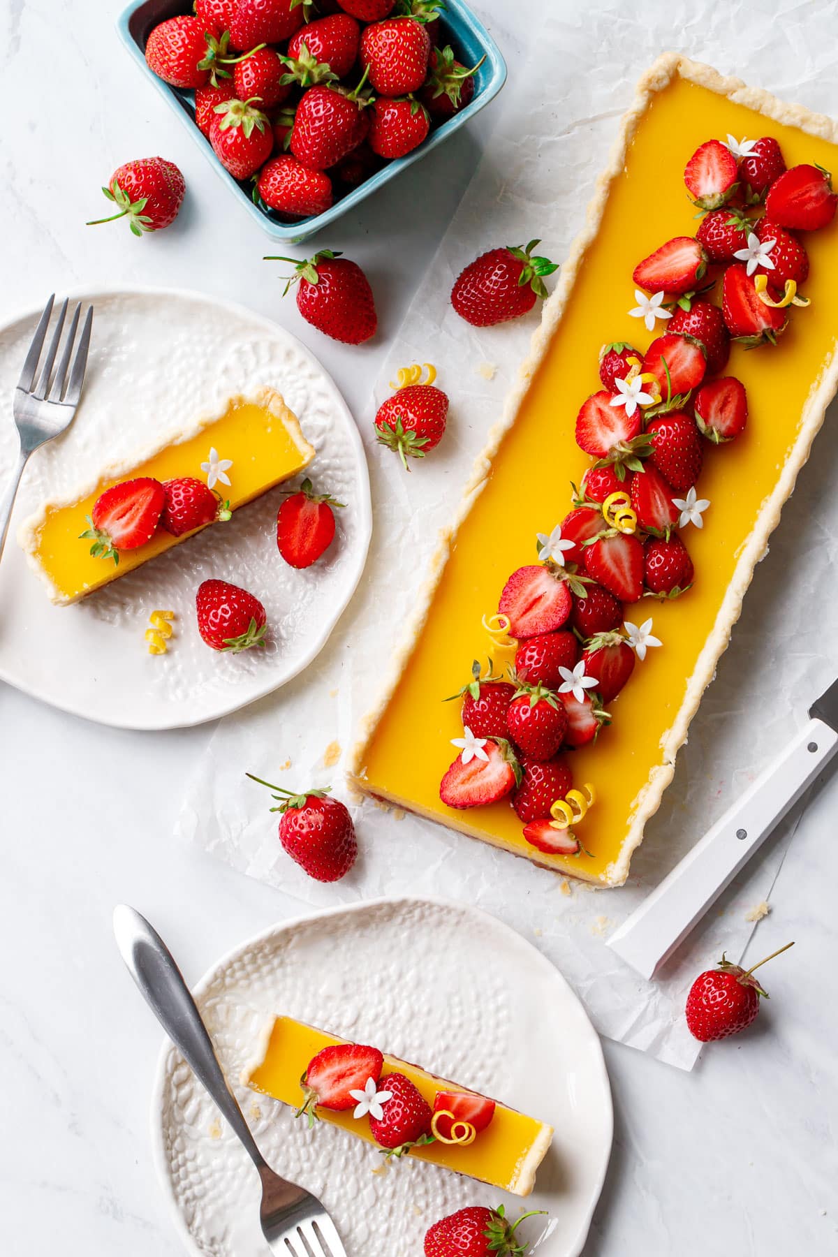 Overhead, plates with slices of Strawberry Meyer Lemon Tart, plus the whole rectangular tart and a basket of strawberries on the side.