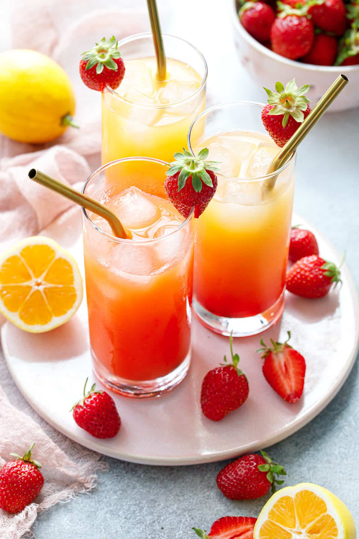 Three glasses of Strawberry Passionfruit Lemonade with metal straws, plus cut lemons and strawberries and a bowl of berries in the background.