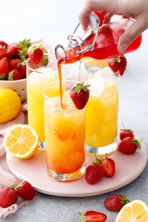 Pouring strawberry syrup into 3 glasses filled with ice and a bright yellow lemon passionfruit juice.