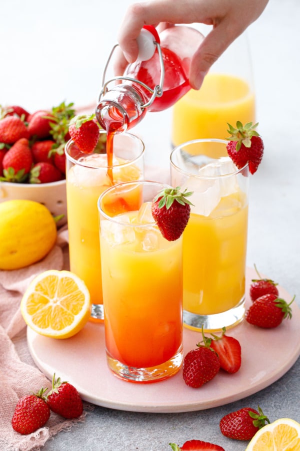 Pouring strawberry syrup into 3 glasses filled with ice and a bright yellow lemon passionfruit juice.