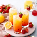 Three glasses with pretty ombre colored Strawberry Passionfruit Lemonade, with black passionfruit seeds speckled throughout and cut lemons and strawberries scattered around.