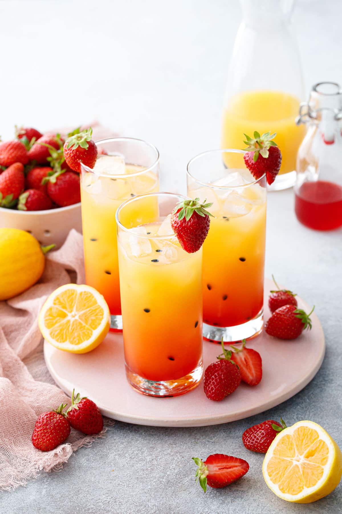 Три стакана с красивым клубничным лимонадом маракуйи цвета омбре, с черными крапинками семян маракуйи и нарезанными лимонами и клубникой, разбросанными вокруг.